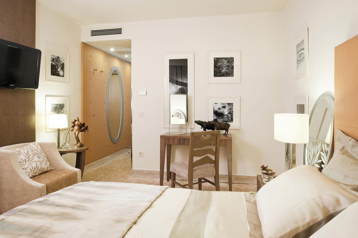 A hotel room with a bed, a desk with a chair, a TV mounted on the wall, and framed pictures on the walls.