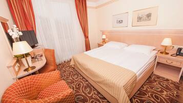 A hotel room with a bed, two bedside tables with lamps, a chair, a desk with a lamp, a mirror, and two framed pictures on the wall.