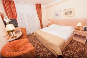 Habitación de hotel con una cama, dos mesitas de noche con lámparas, una silla, un escritorio con lámpara, un espejo y dos cuadros en la pared.