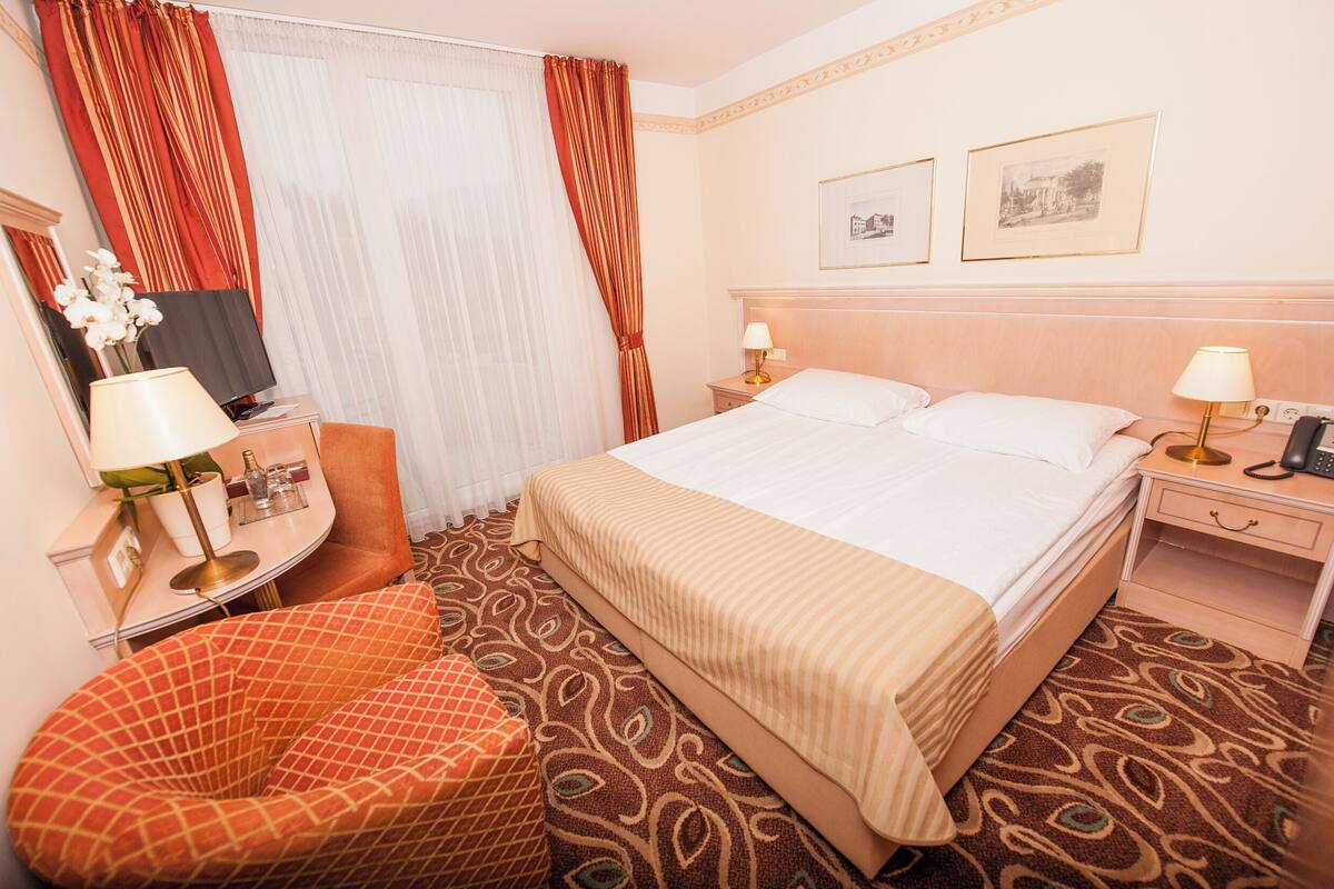 A hotel room with a bed, two bedside tables with lamps, a chair, a desk with a lamp, a mirror, and two framed pictures on the wall.