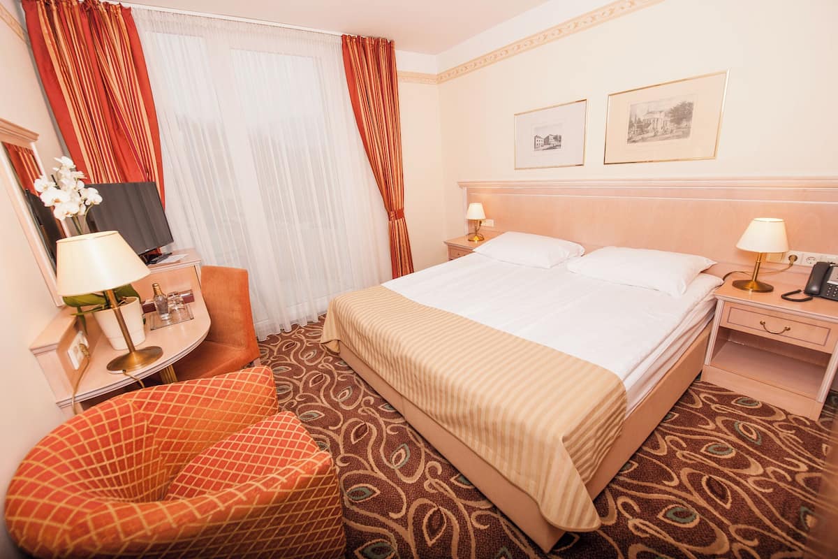 Une chambre d’hôtel comprenant un lit, deux tables de chevet avec des lampes, une chaise, un bureau avec une lampe, un miroir et deux tableaux encadrés accrochés au mur.