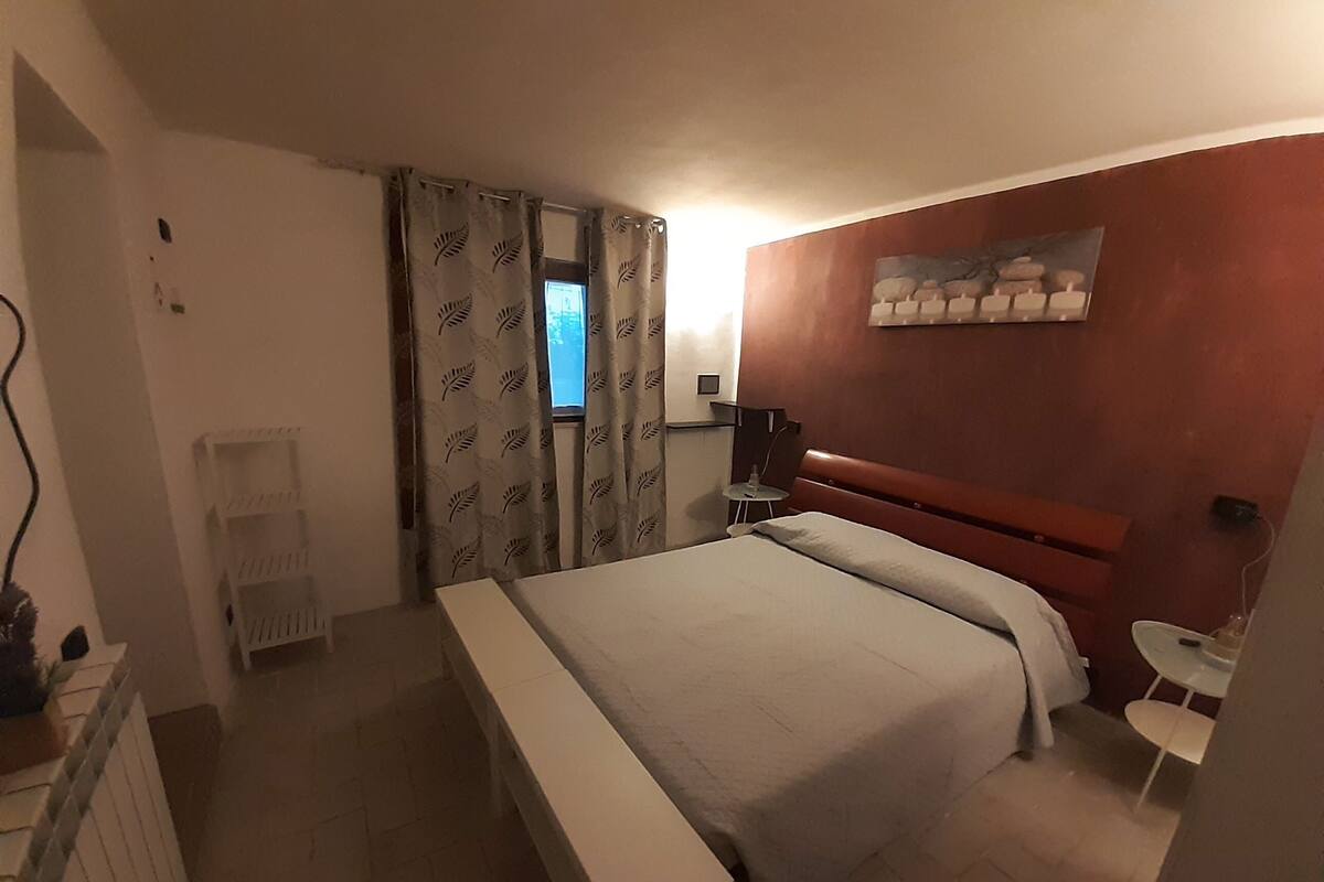 A hotel room with a bed, a small bedside table, a chair, a window with curtains, and a wall-mounted artwork.