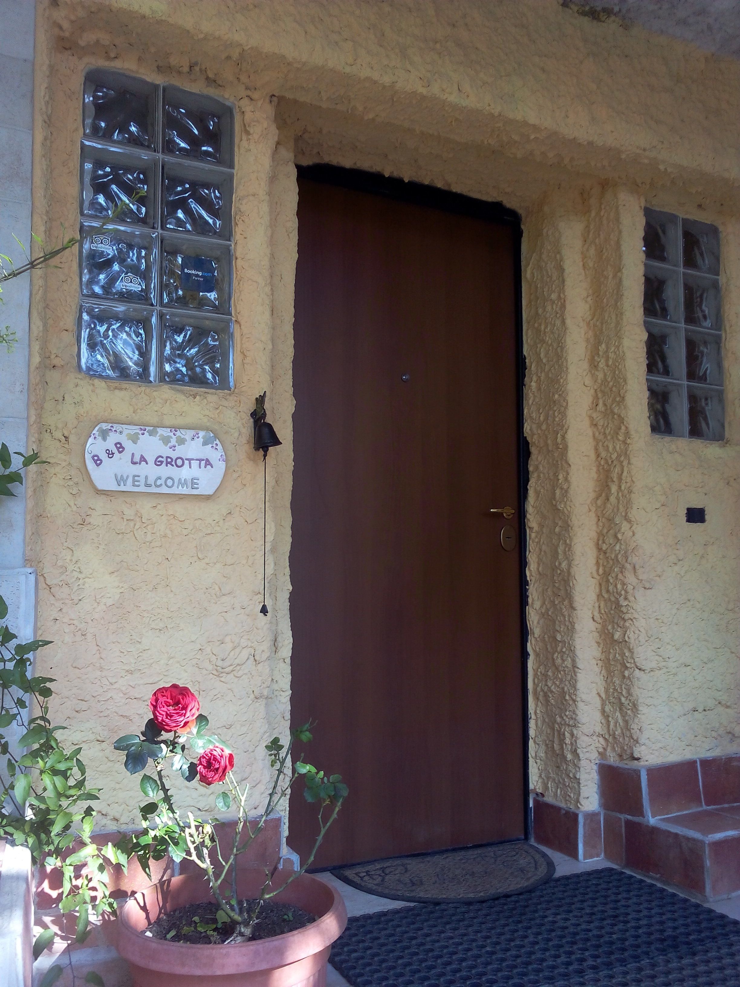 Entrada de un edificio con una puerta de madera, un letrero de bienvenida y una planta en maceta con una flor roja.