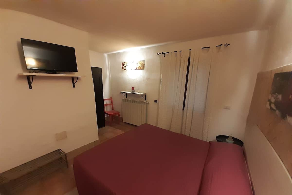 A bedroom with a bed, a TV mounted on the wall, a radiator, a chair, and a door leading to another room.