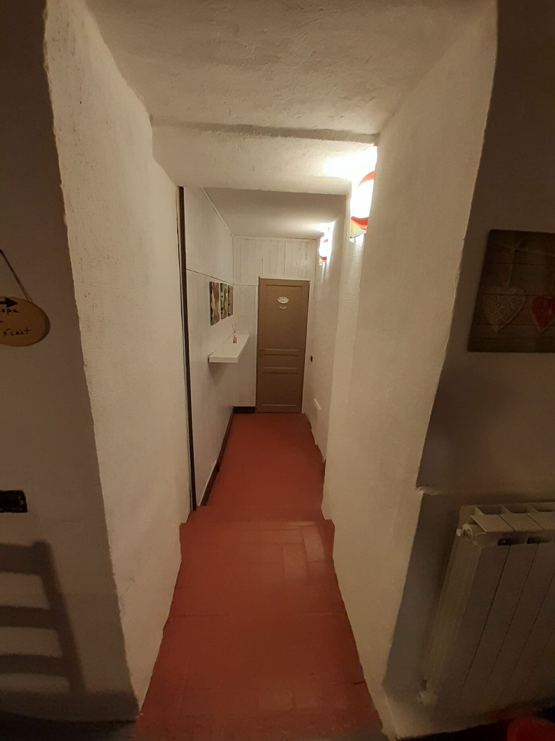Un pasillo estrecho con paredes blancas, una alfombra roja, una puerta al final y un radiador a la derecha.