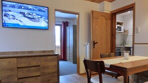 A room with a wooden dining table, a television mounted on the wall displaying a snowy landscape, and a kitchen area visible through an open door. - Aparthotel Buchauer Tirol (Thiersee)