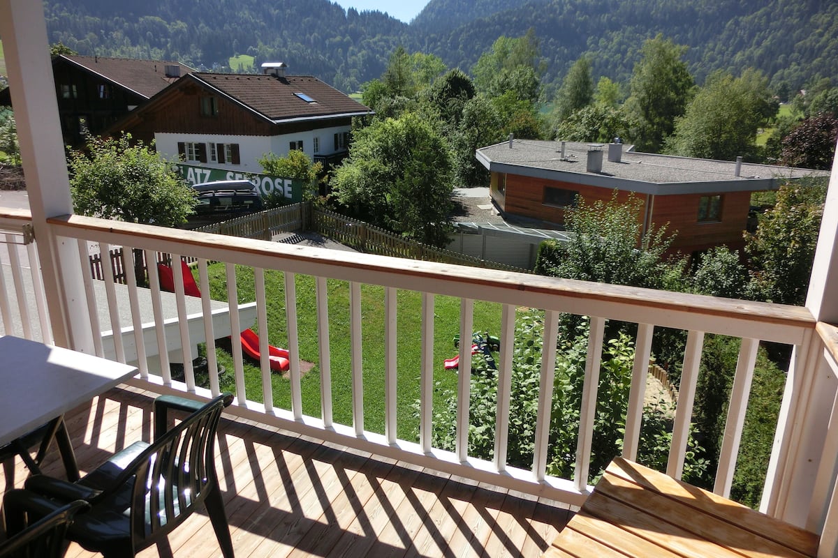 Una terraza de madera con barandilla blanca, con vistas a un valle con casas y vegetación.