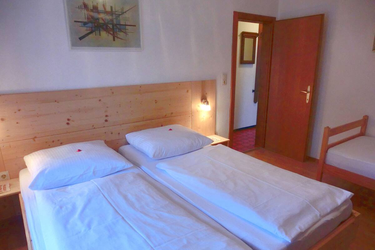 Una habitación con dos camas individuales, un cabecero de madera, una mesita de noche con lámpara y una puerta que da a otra habitación.