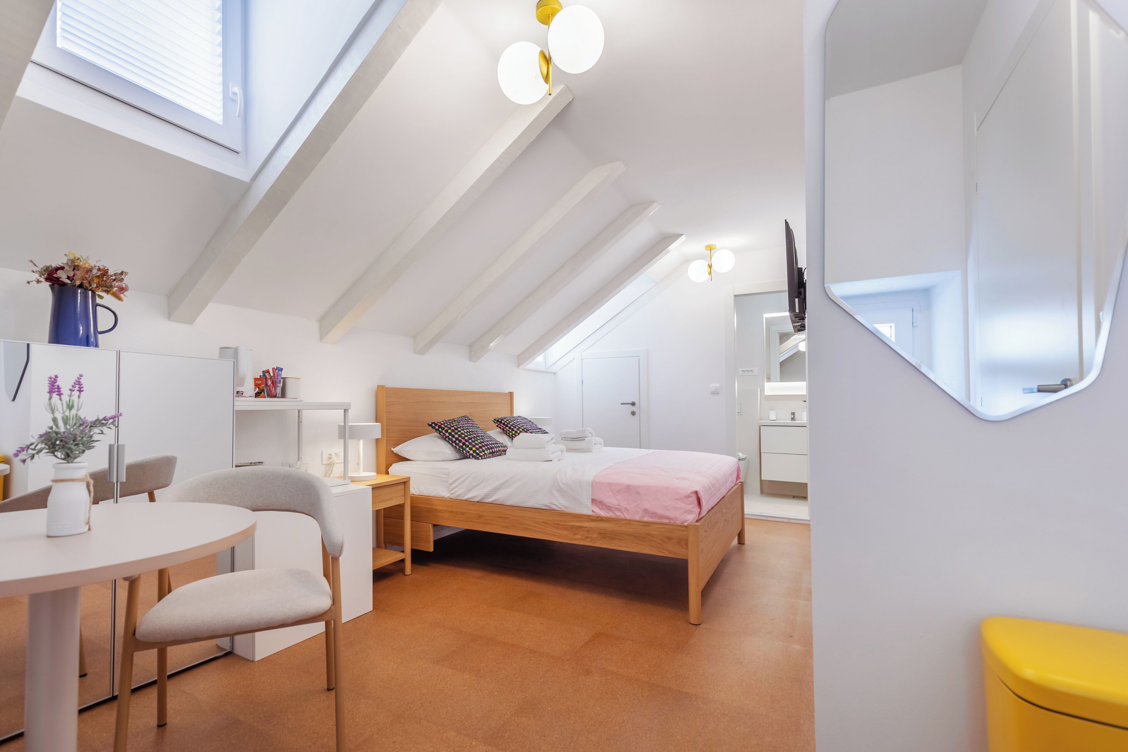 A modern bedroom with a skylight, a bed with pillows, a bedside table, a chair, and a desk with a vase of flowers.