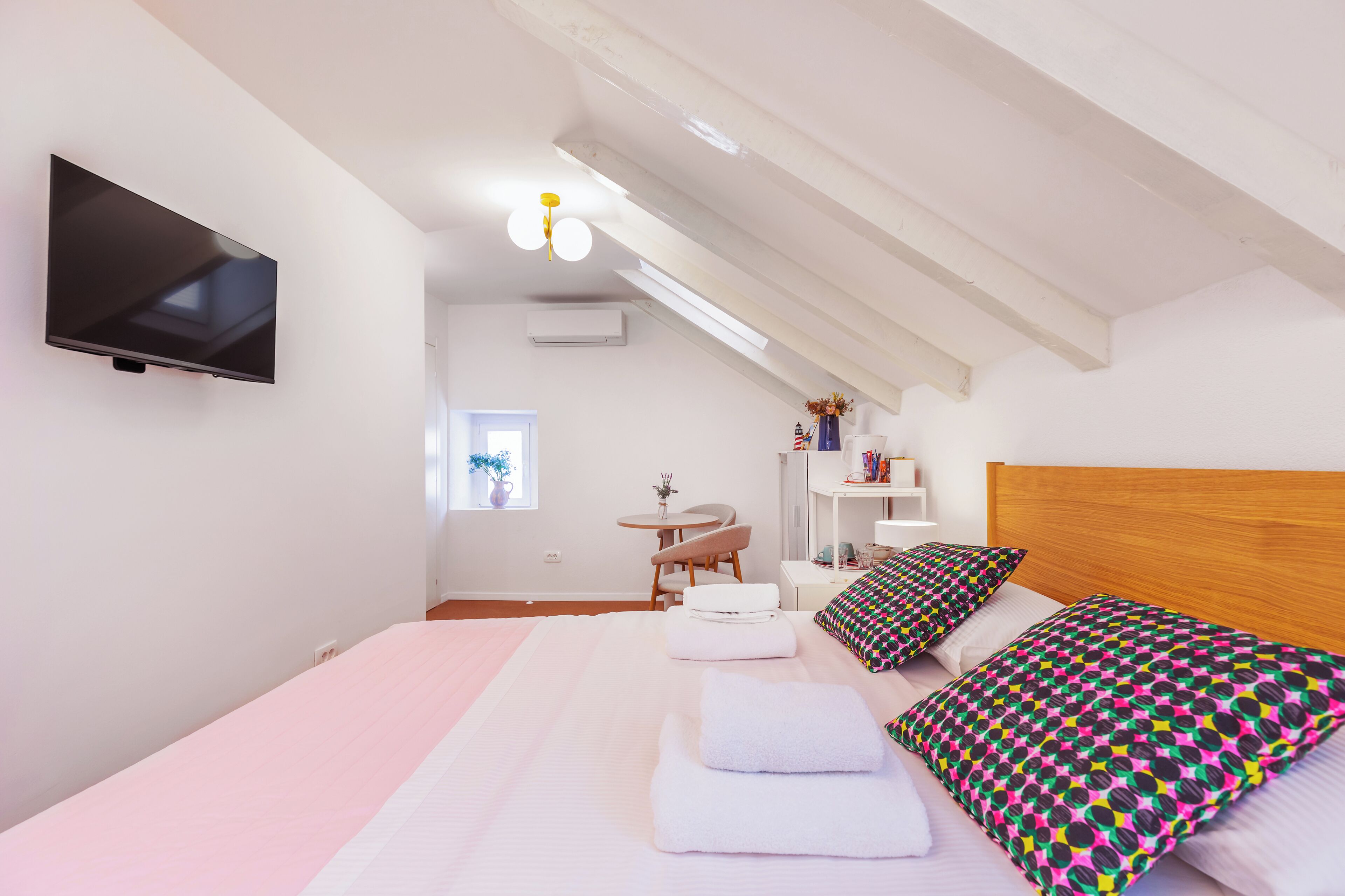 A modern bedroom with a large bed, a TV mounted on the wall, a small table, and a window.