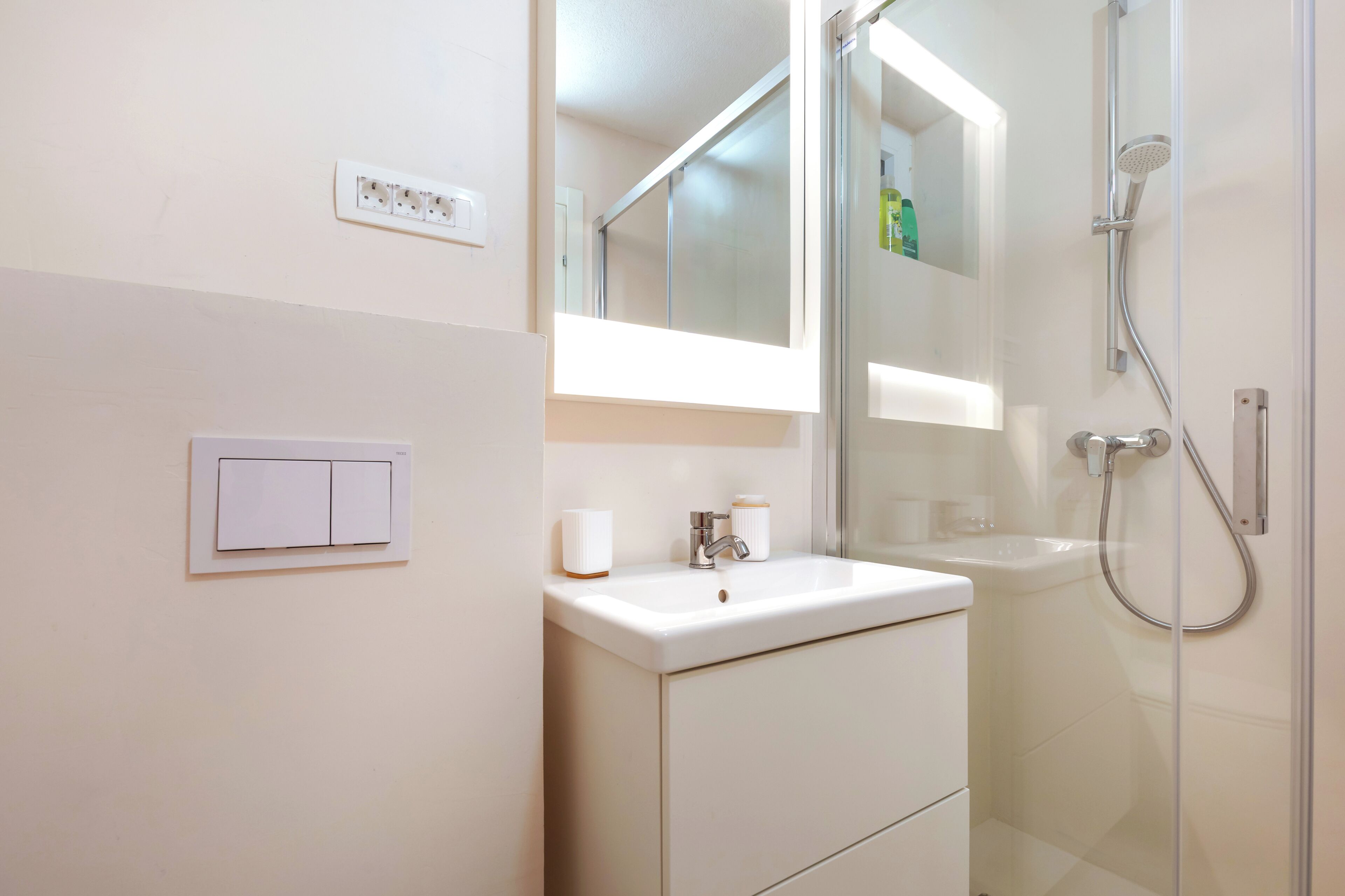 A bathroom with a white sink, mirror, and shower. There is a light switch and a small bottle on the sink.