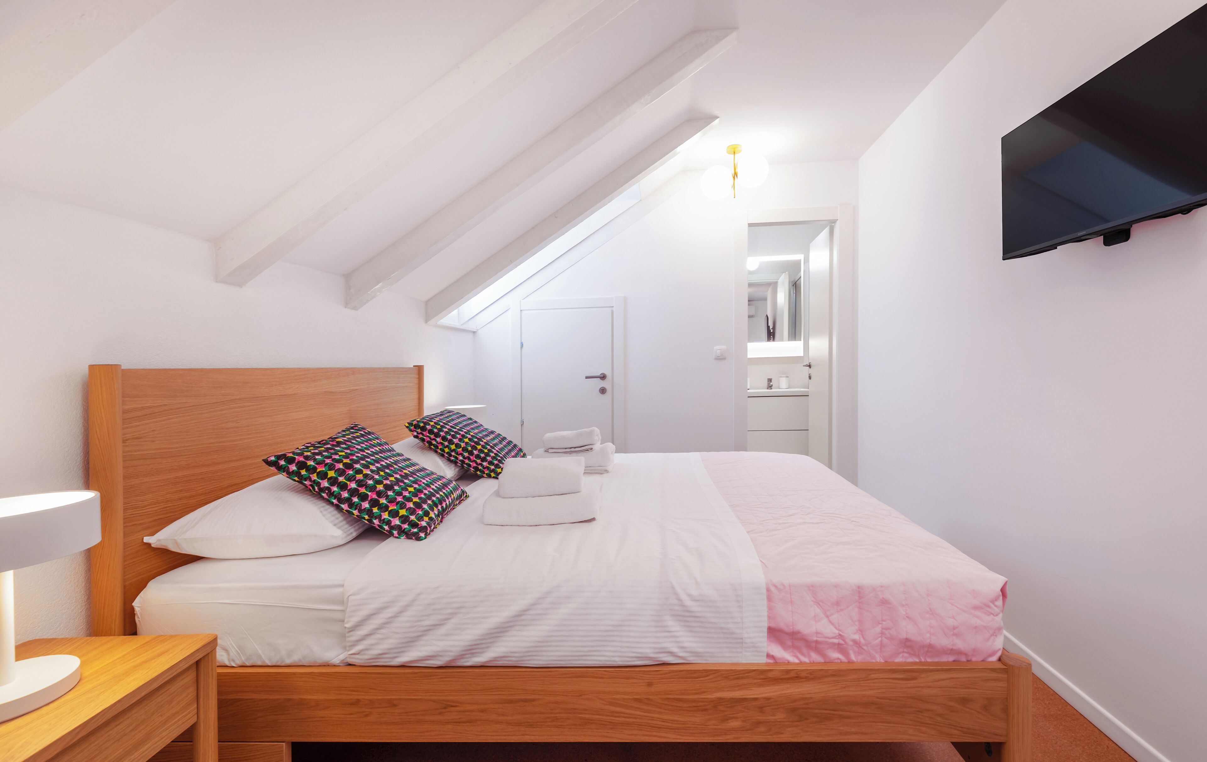 A bedroom with a wooden bed, white bedding, a bedside lamp, a TV on the wall, and a skylight.