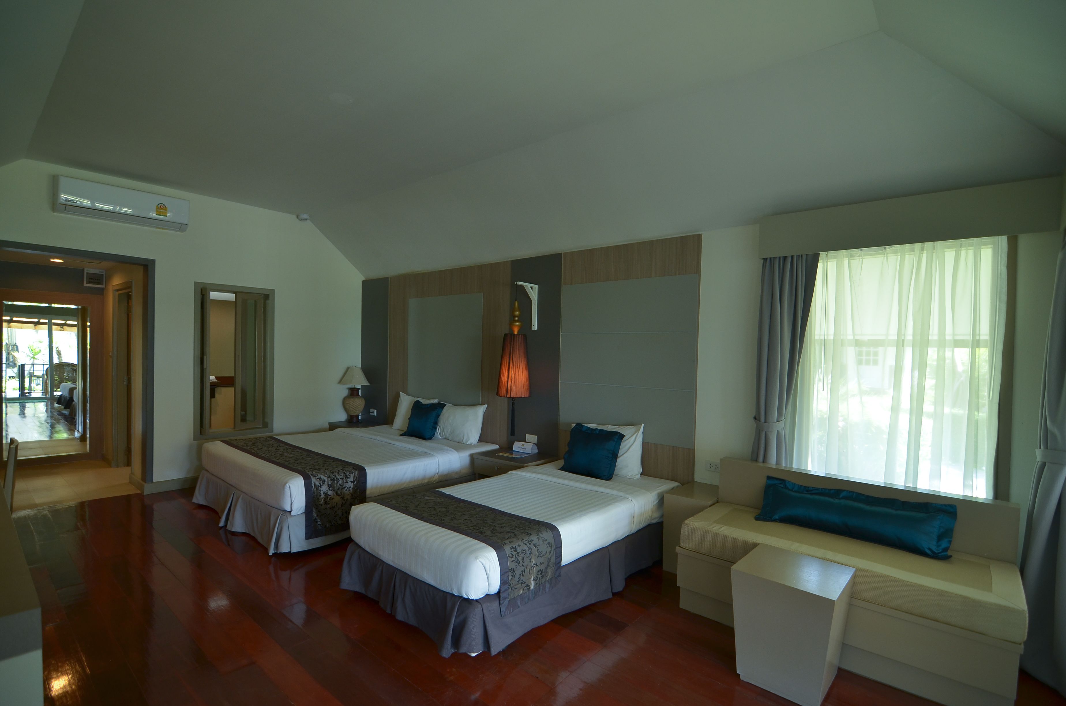 Une chambre d’hôtel avec deux lits, un canapé et une petite table. Il y a une fenêtre avec des rideaux, une lampe et un appareil de climatisation.