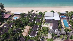 An aerial view of a resort with multiple bungalow-style buildings, a central swimming pool, and a beach in the background.