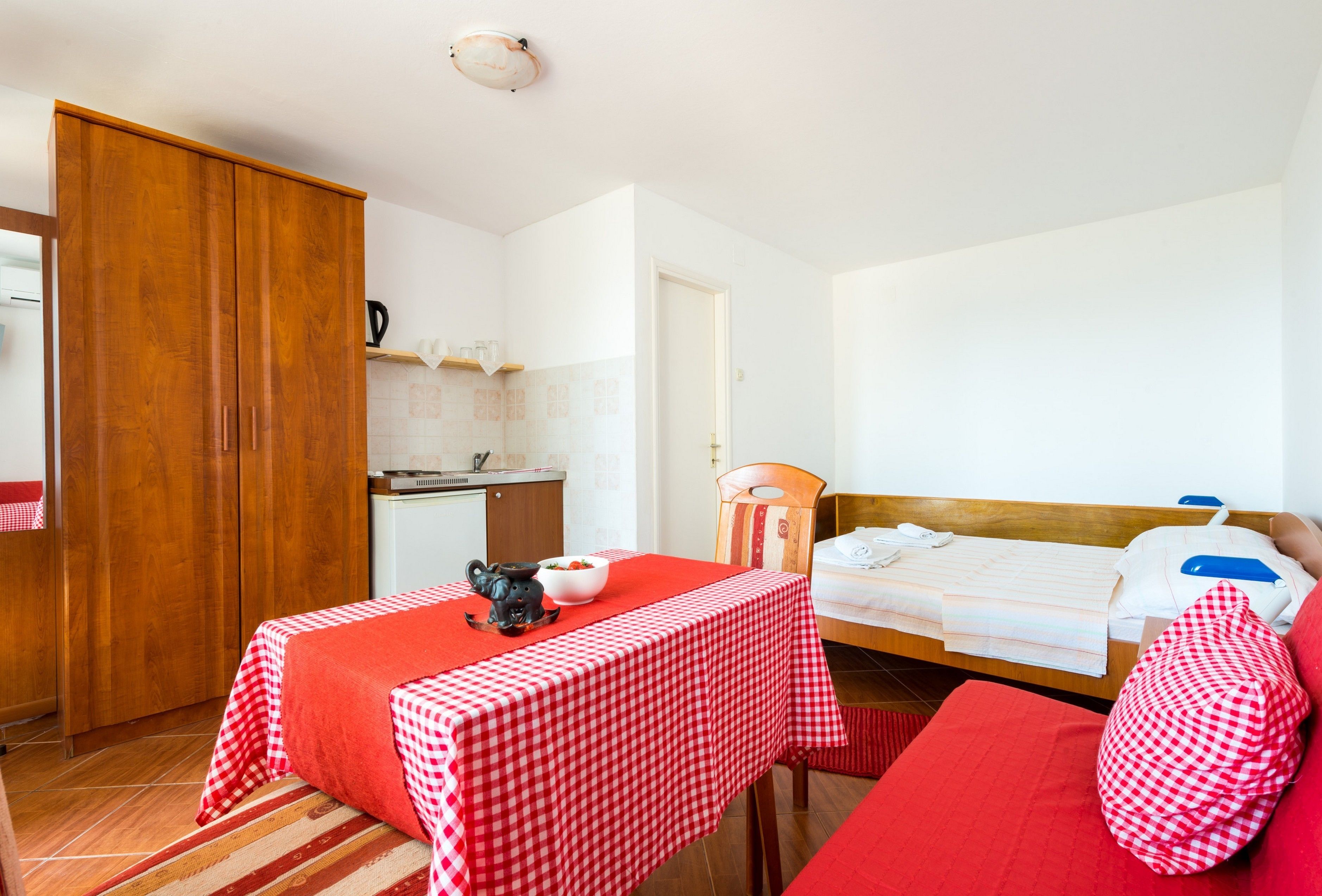 Una habitación con cama, una mesa con mantel a cuadros rojos, un armario de madera y una kitchenette.