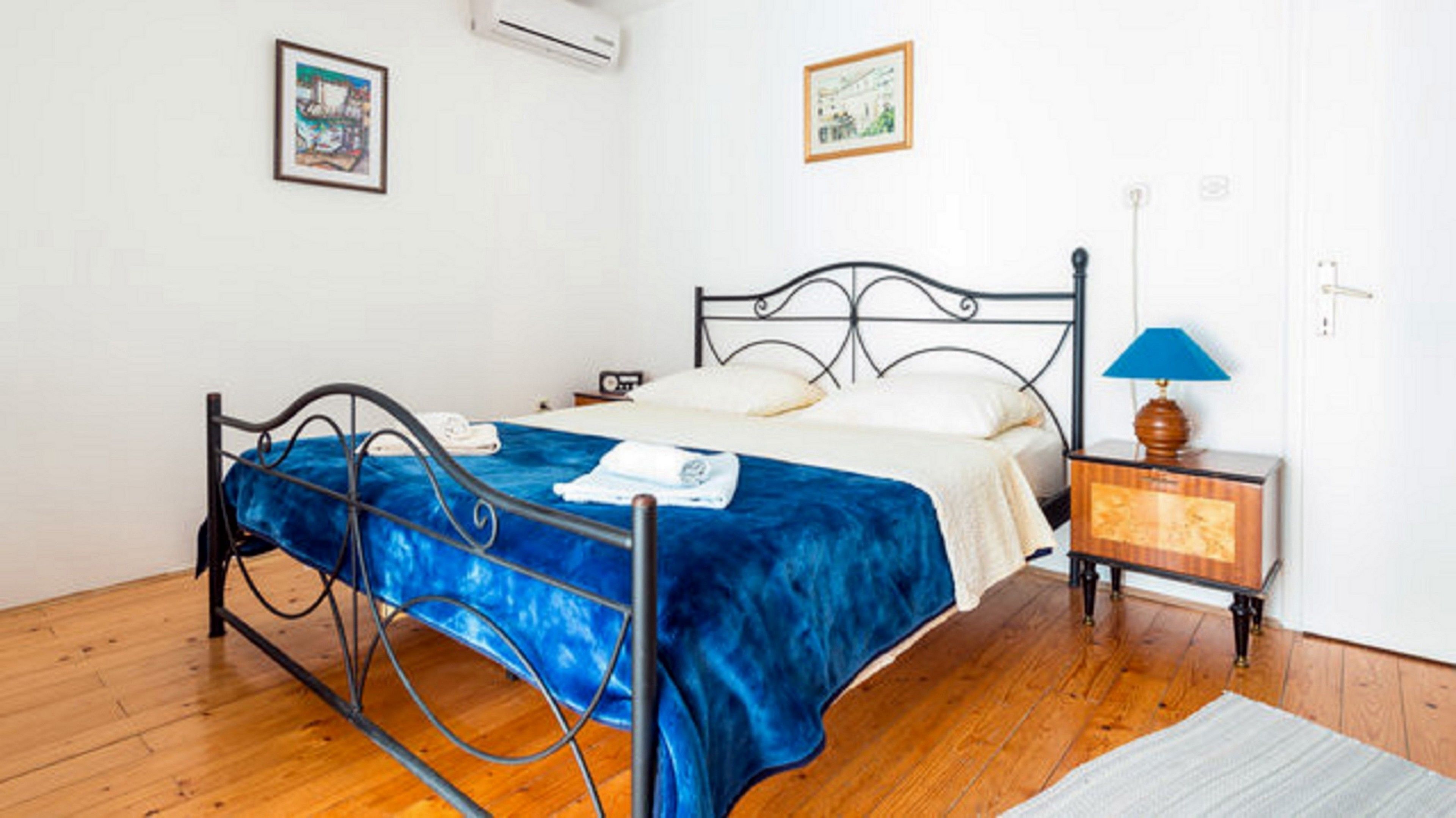 Een slaapkamer met een metalen bedframe, een blauw bedlaken, een nachtkastje met een lamp en twee ingelijste foto's aan de muur.