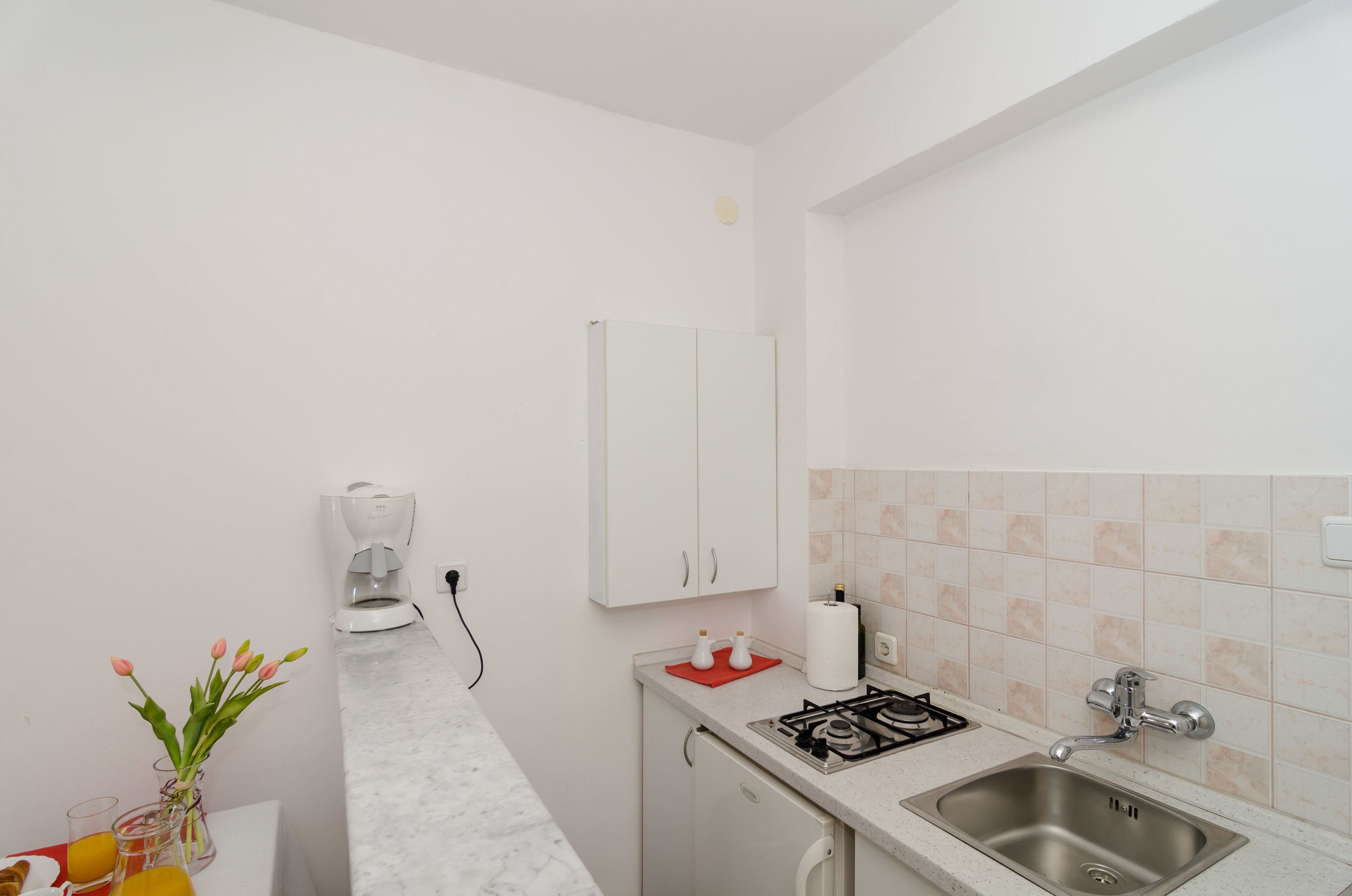 A small kitchenette with a gas stove, sink, and white cabinets. There is a coffee maker and a vase with flowers on the counter.