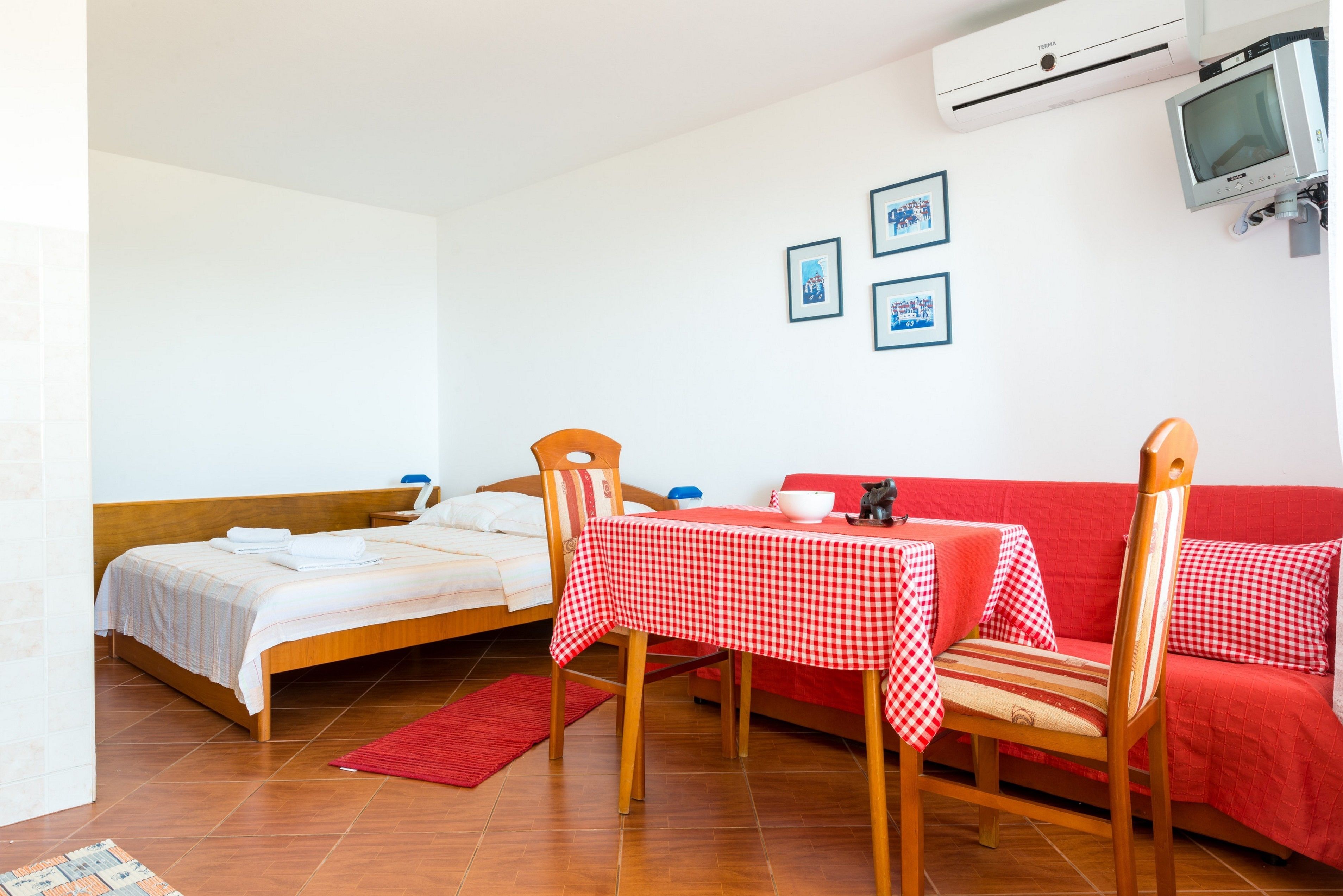 A room with a bed, a red sofa, a table with a checkered tablecloth, and a television mounted on the wall.
