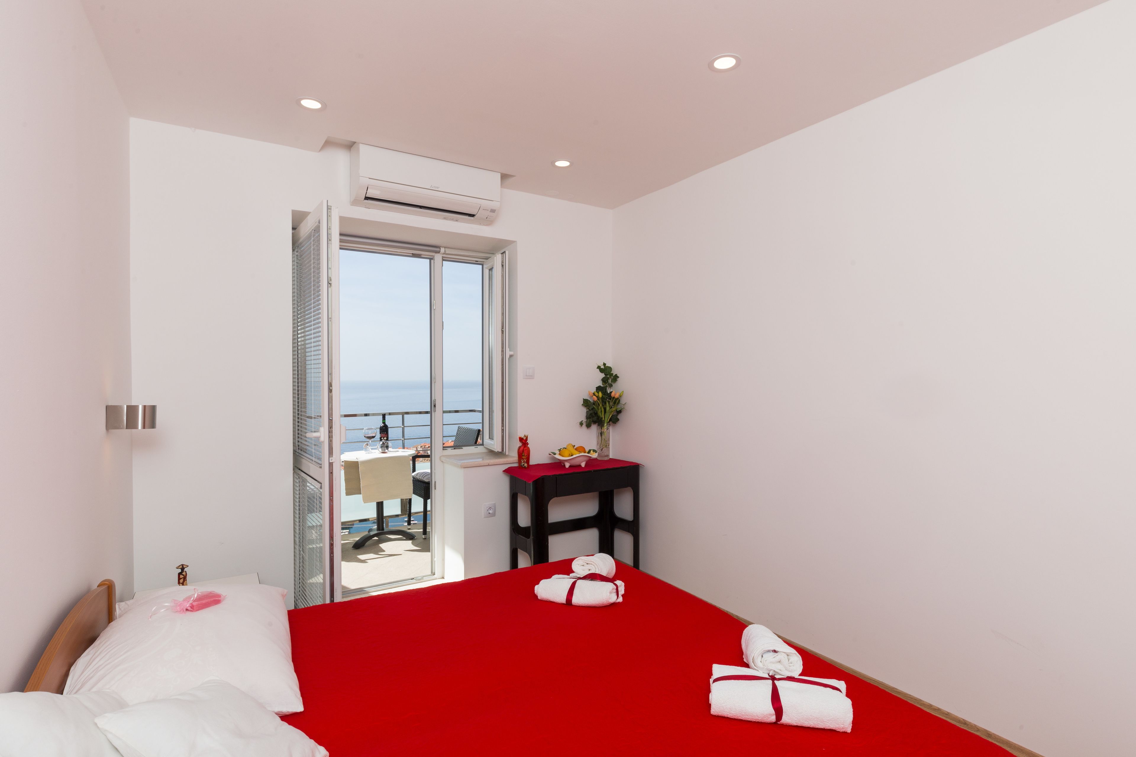 A hotel room with a red bedspread, white pillows, a small table with a vase and fruit, and a balcony with a view.