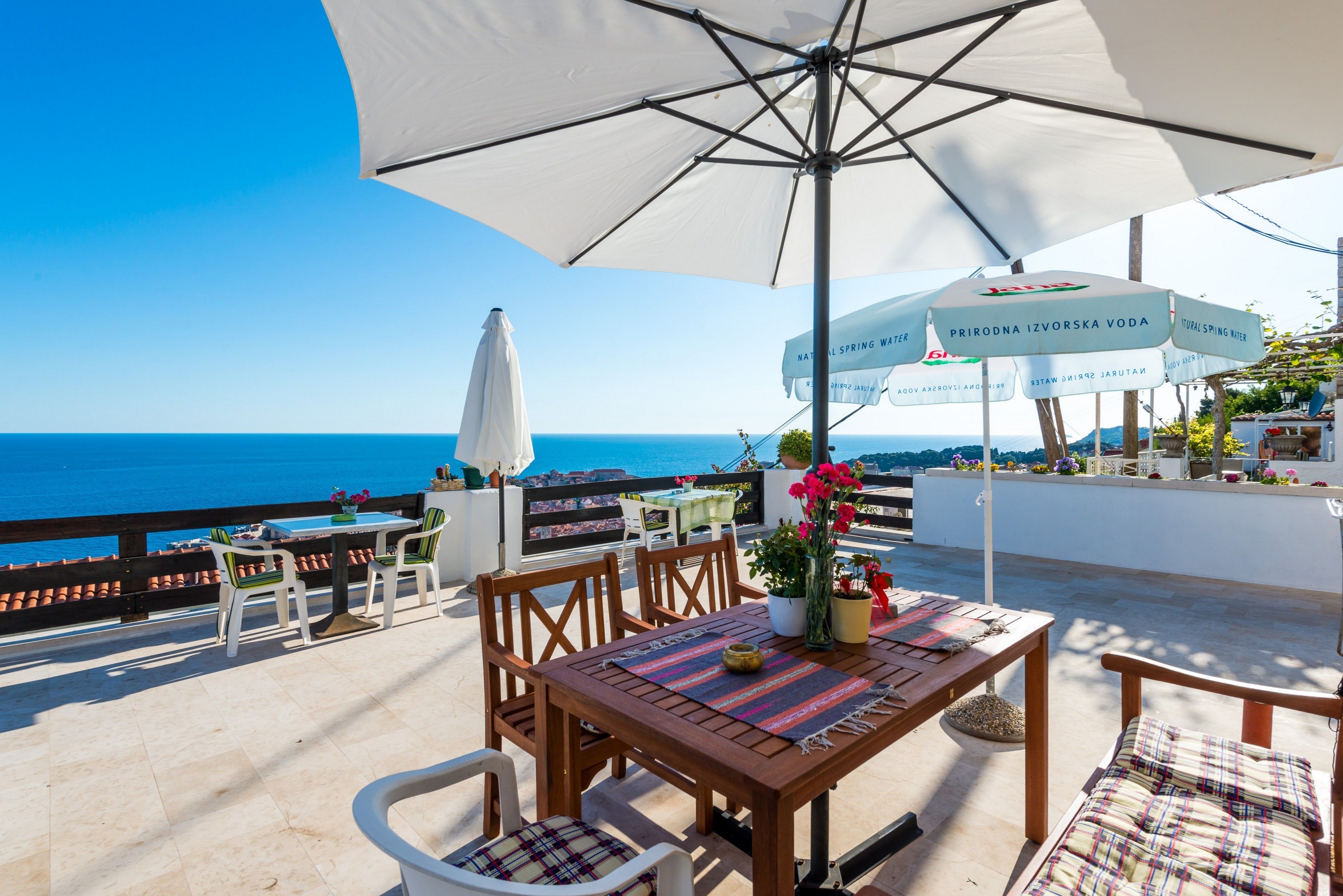 Una terraza con una mesa de madera, sillas y un paraguas grande. Hay plantas en macetas y vistas al mar.