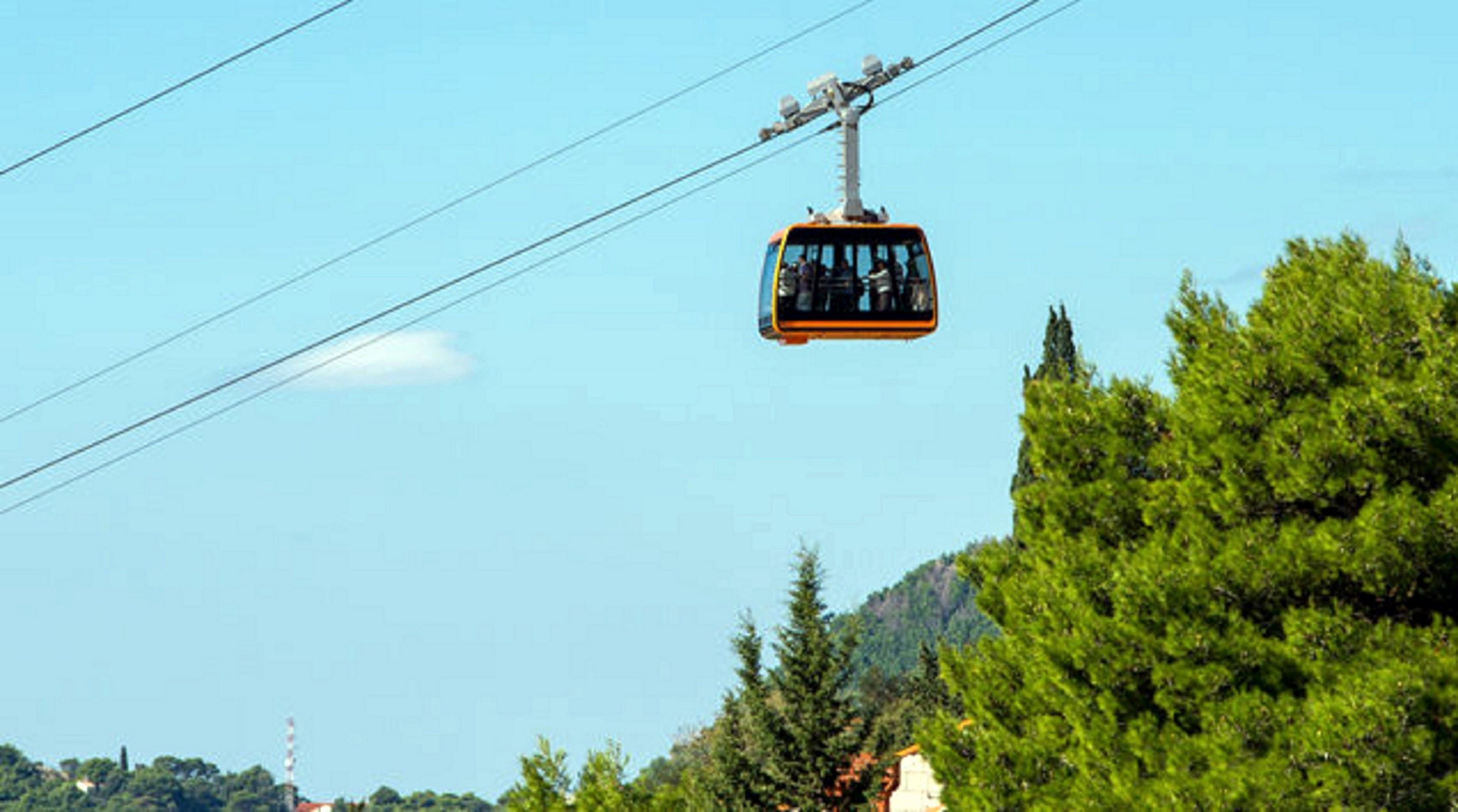 A cable car in motion against a clear sky, with dense green trees and a distant building visible.
