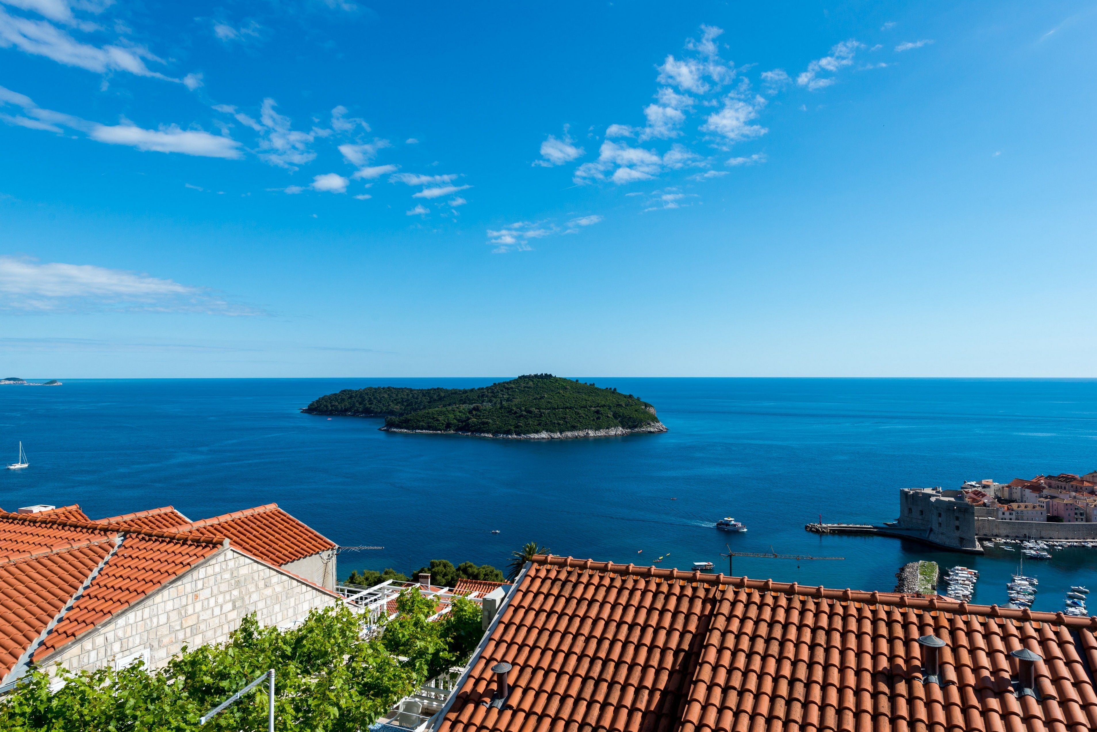 A coastal town with terracotta-roofed buildings, a small island, and clear blue skies.