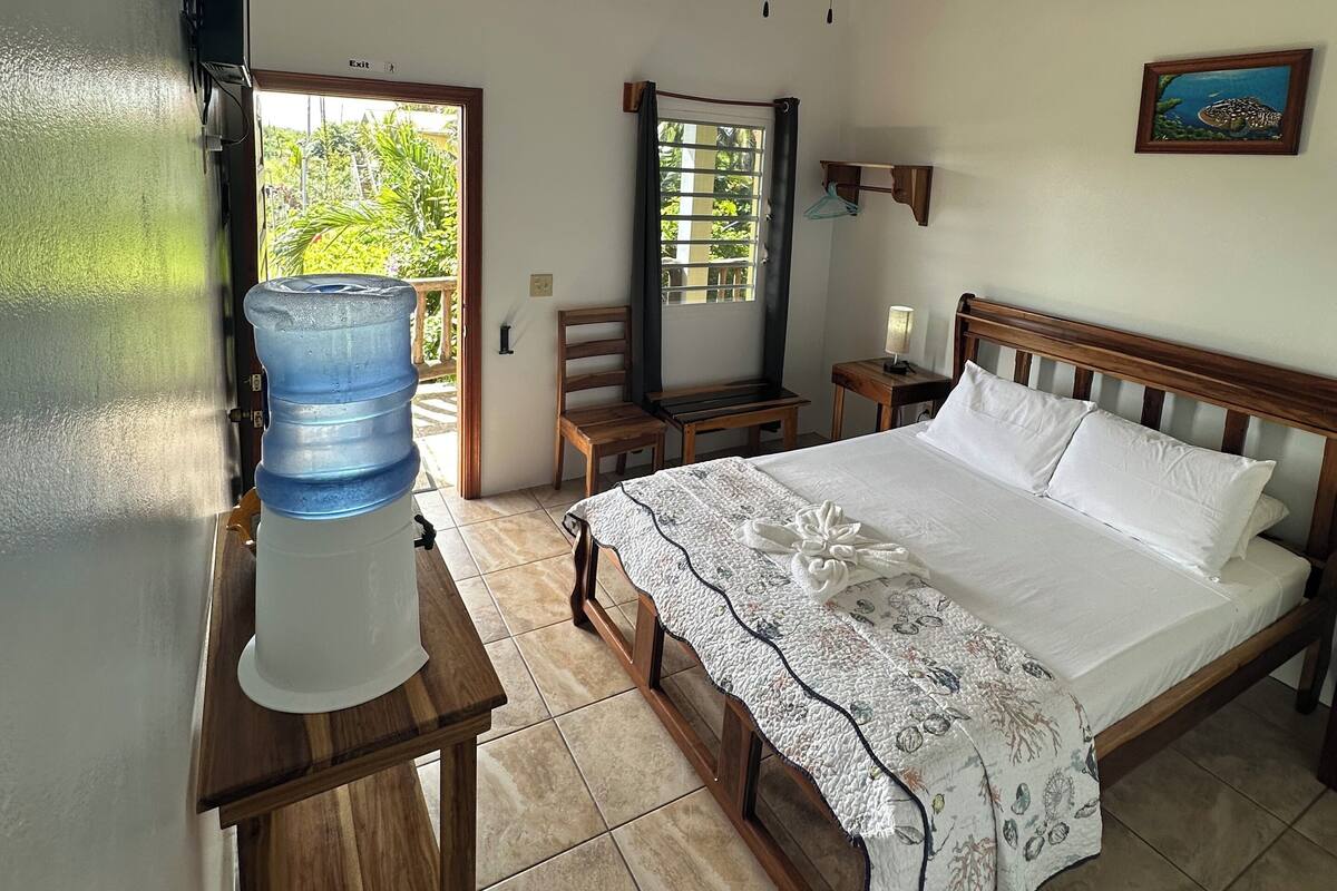 A bedroom with a bed, a ceiling fan, a water dispenser, a wooden headboard, a nightstand, and a painting on the wall.