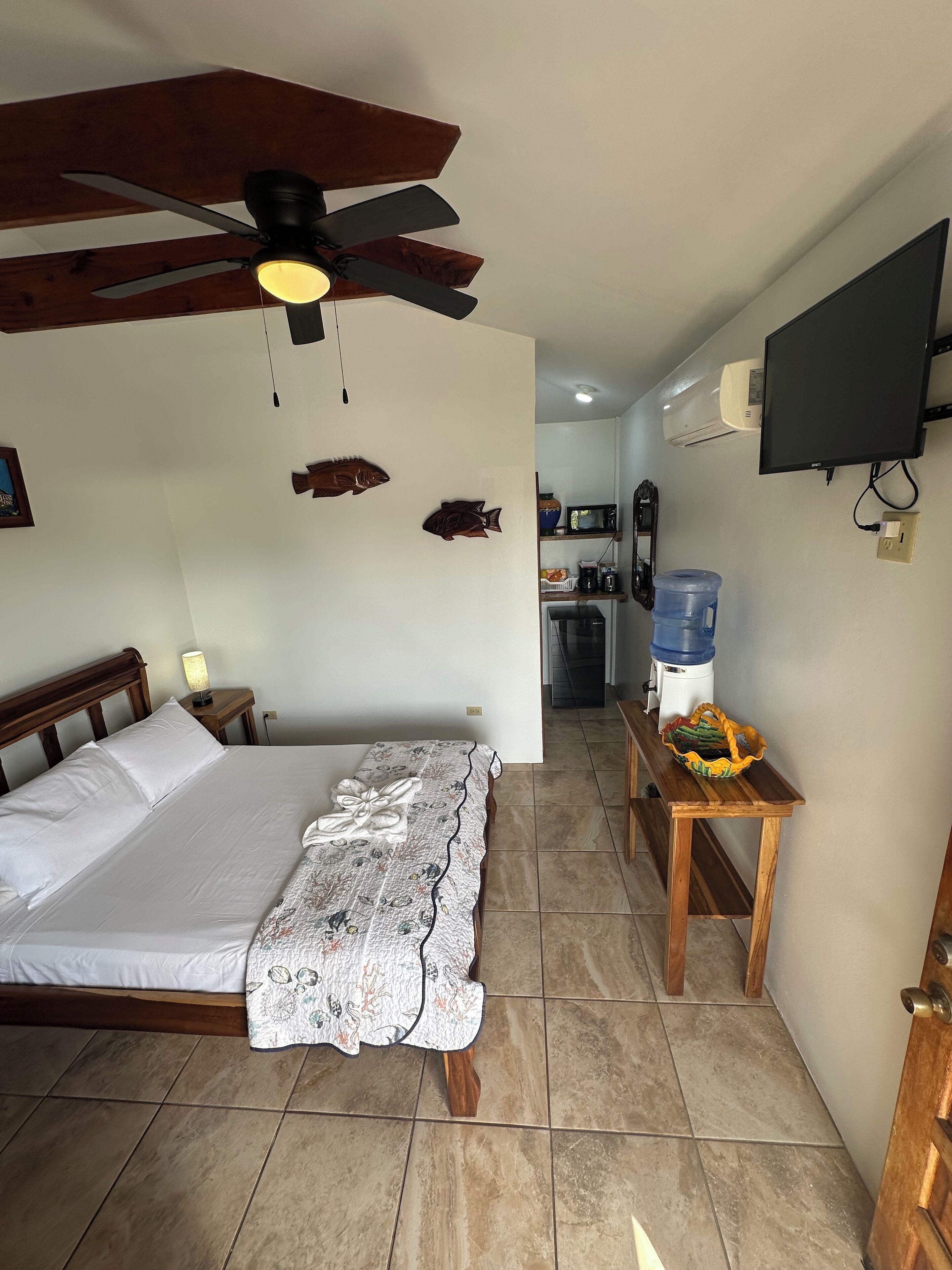 Una habitación de hotel con cama, ventilador de techo, dispensador de agua y televisión montada en la pared.