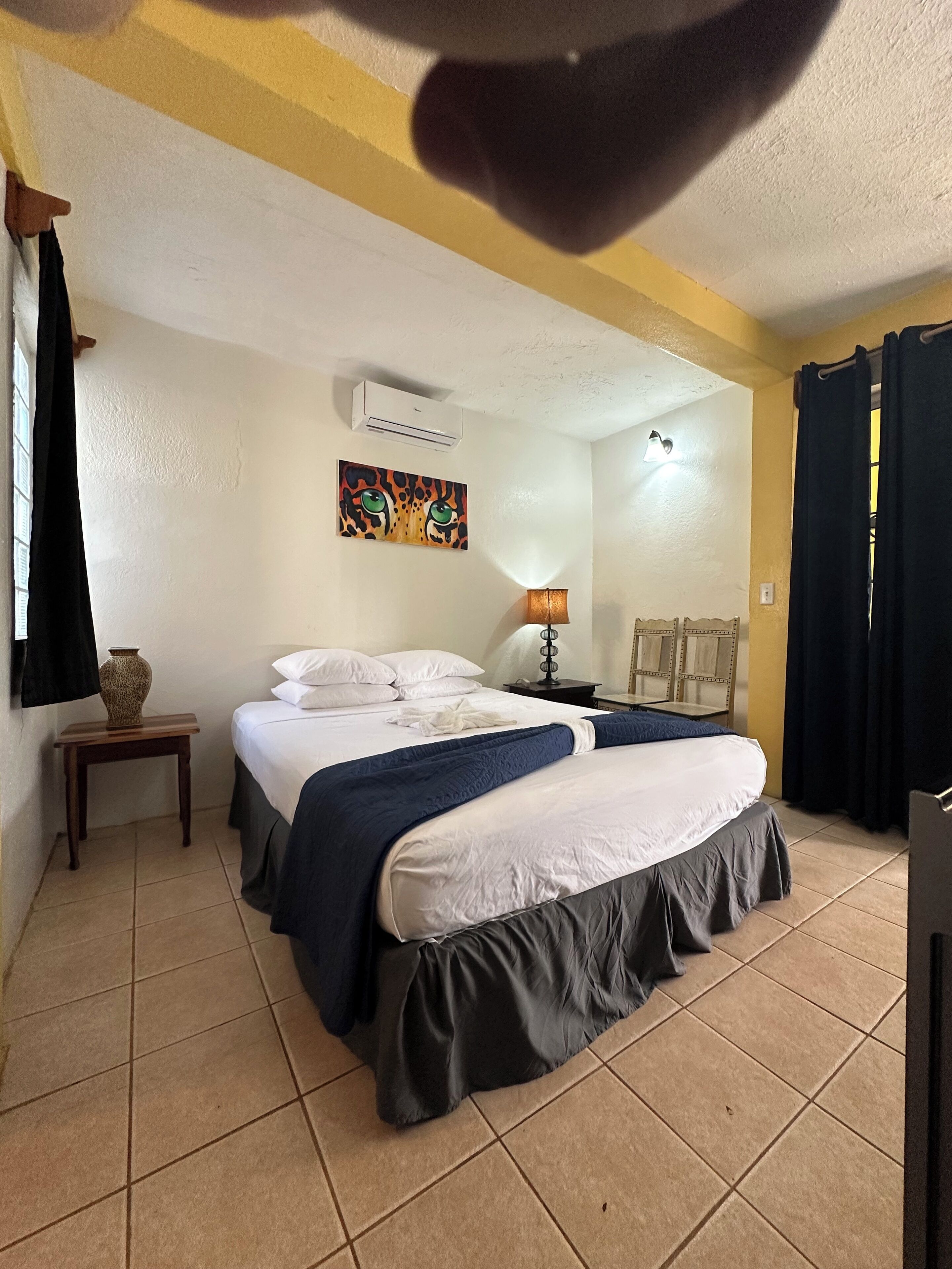 Una habitación de hotel con una cama, una mesita de noche, una lámpara, un cuadro y una ventana con cortinas.
