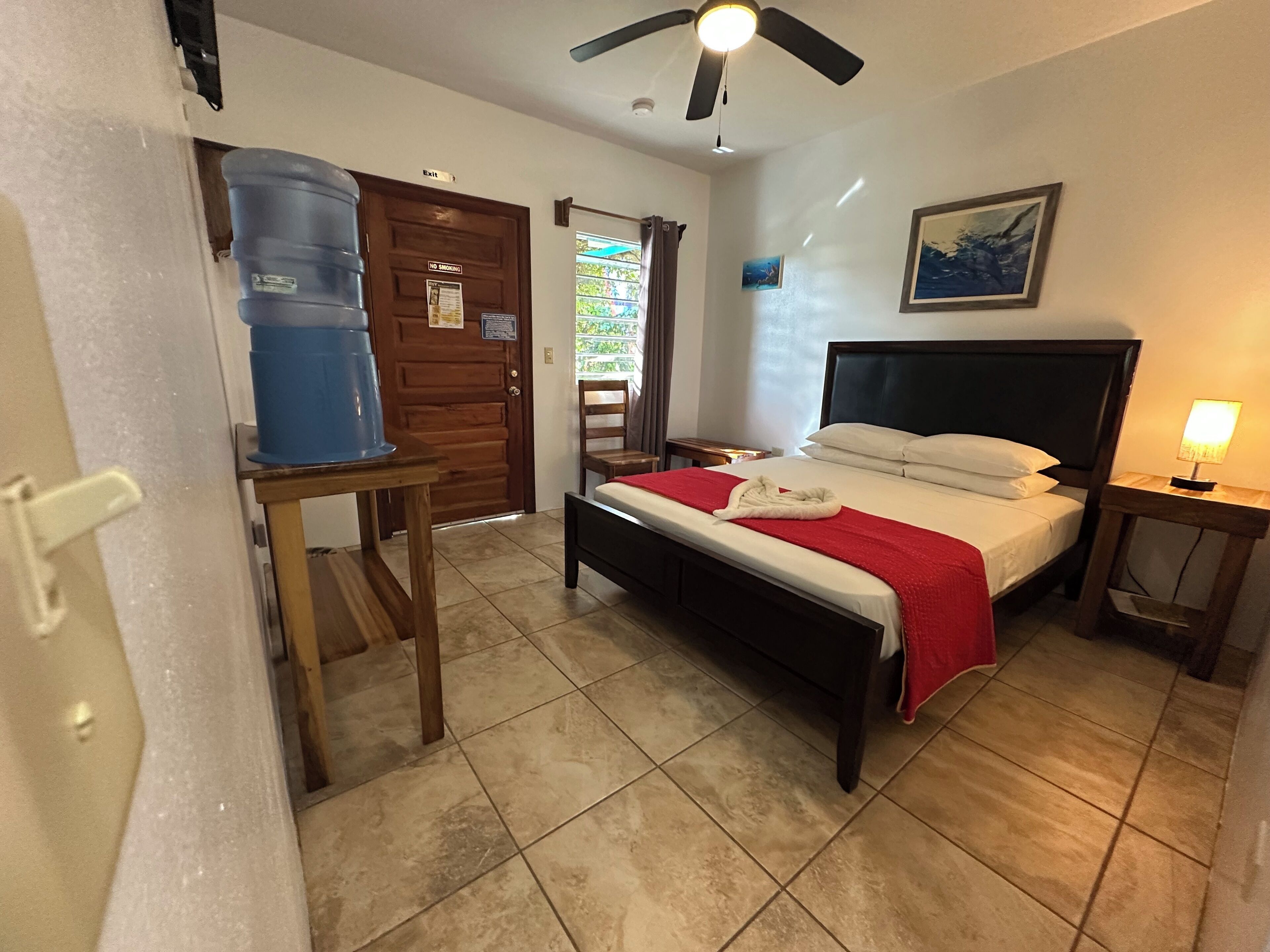 Habitación de hotel con cama, mesita de noche, escritorio con dispensador de agua, ventilador de techo y ventana con cortinas.