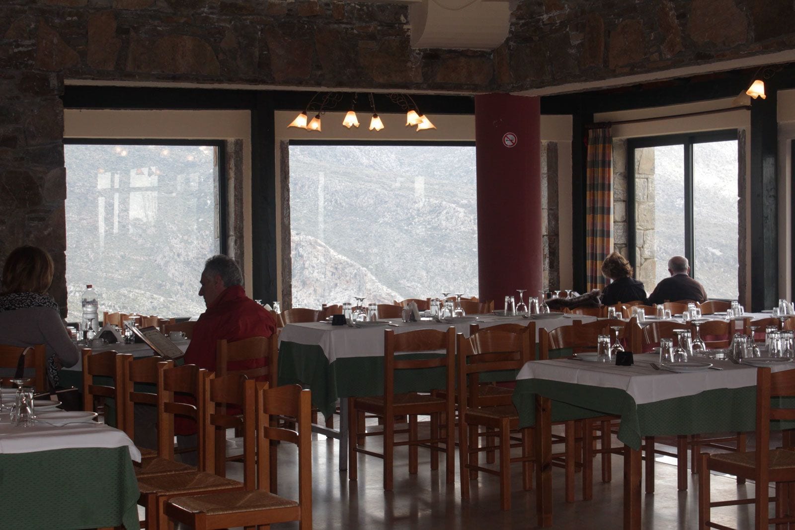 A restaurant with multiple tables and chairs, large windows with a mountain view, and a stone wall interior.