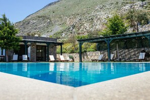 Ein Pool mit Liegestühlen und einem Pavillon, umgeben von einer Steinmauer und einem Bergpanorama.
