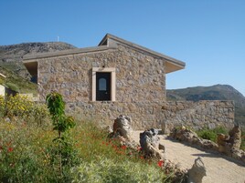 Edificio de piedra con una pequeña ventana y puerta, rodeado de vegetación natural y un camino de tierra.