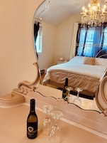 Un dormitorio con un espejo grande, una cama, una mesita de noche con una botella de vino y copas, y un candelabro.