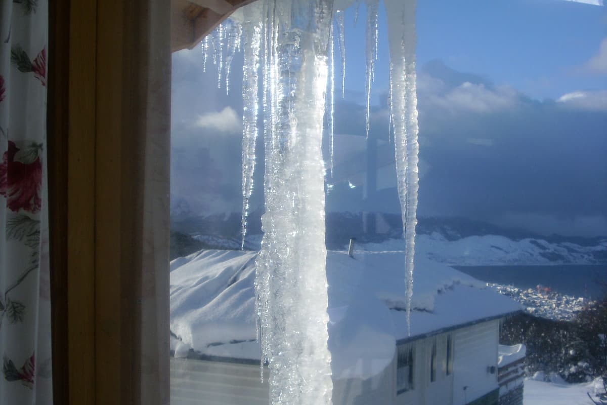 Des glaçons pendent d’une fenêtre, un toit recouvert de neige et un paysage enneigé avec des montagnes en arrière-plan.