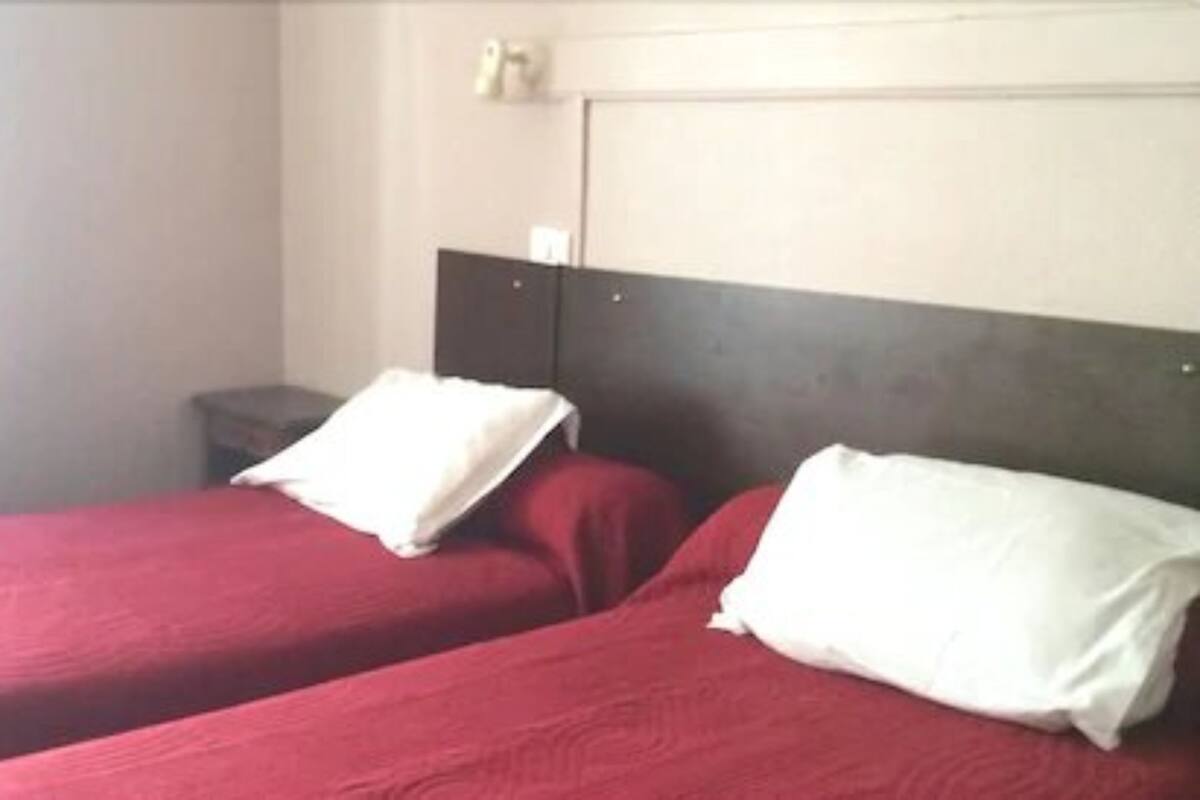 Habitación de hotel con dos camas individuales, cada una con una almohada blanca y una colcha roja. Hay una mesita de noche pequeña con una lámpara.