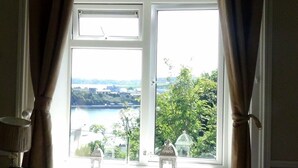 A room with a large window, curtains, a lamp, and a view of a lake and greenery. - Robin Hill House Heritage B&B (Cobh)