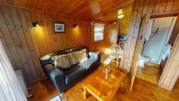 A cozy cabin interior with a wooden ceiling, a black leather sofa with cushions, a wooden coffee table, and a white chair.