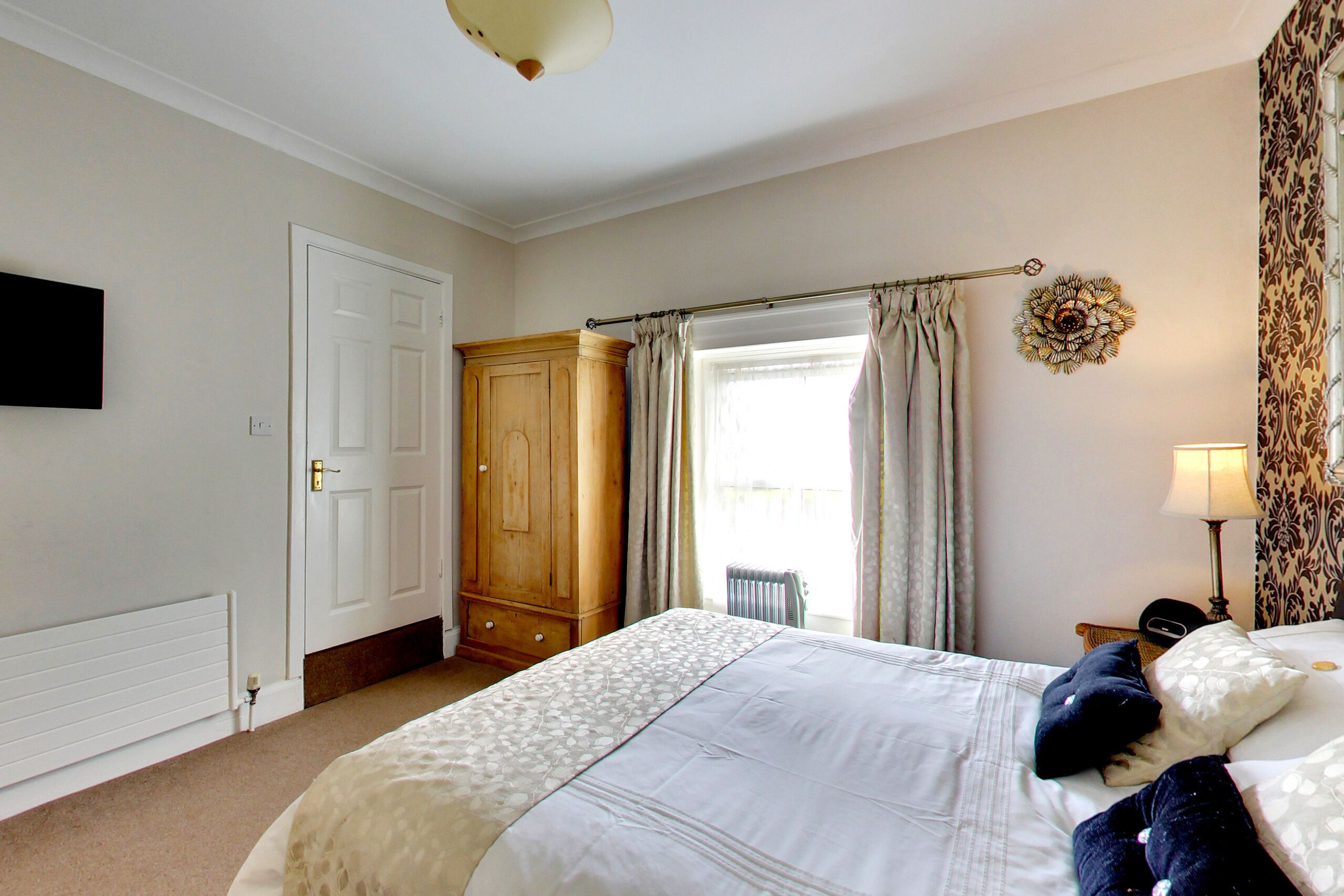 A bedroom with a bed, a wardrobe, a radiator, a window with curtains, a wall-mounted TV, and a lamp.