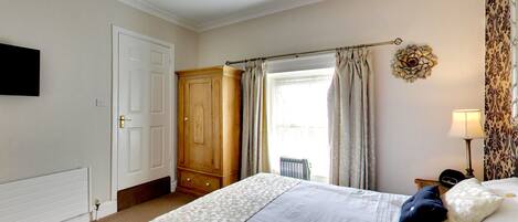 A bedroom with a bed, a wardrobe, a radiator, a window with curtains, a wall-mounted TV, and a lamp.