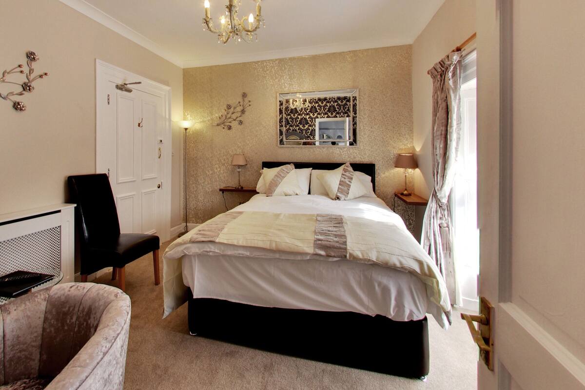 A bedroom with a large bed, a chair, a desk, and a mirror.