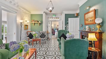 A hotel lobby with a checkered floor, green upholstered chairs, a chandelier, and a fireplace with a lamp.