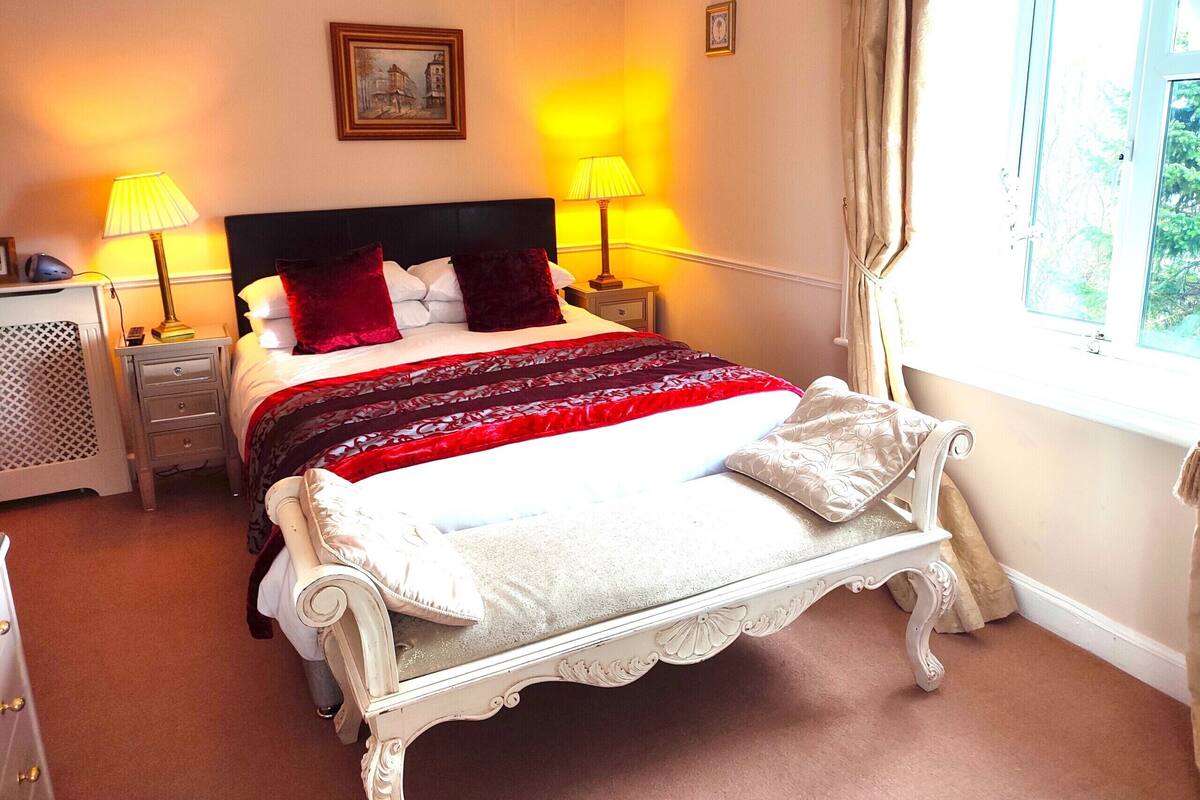 A neatly made bed with a red and white bedspread, two bedside lamps, a nightstand, a framed picture on the wall, and a window with curtains.