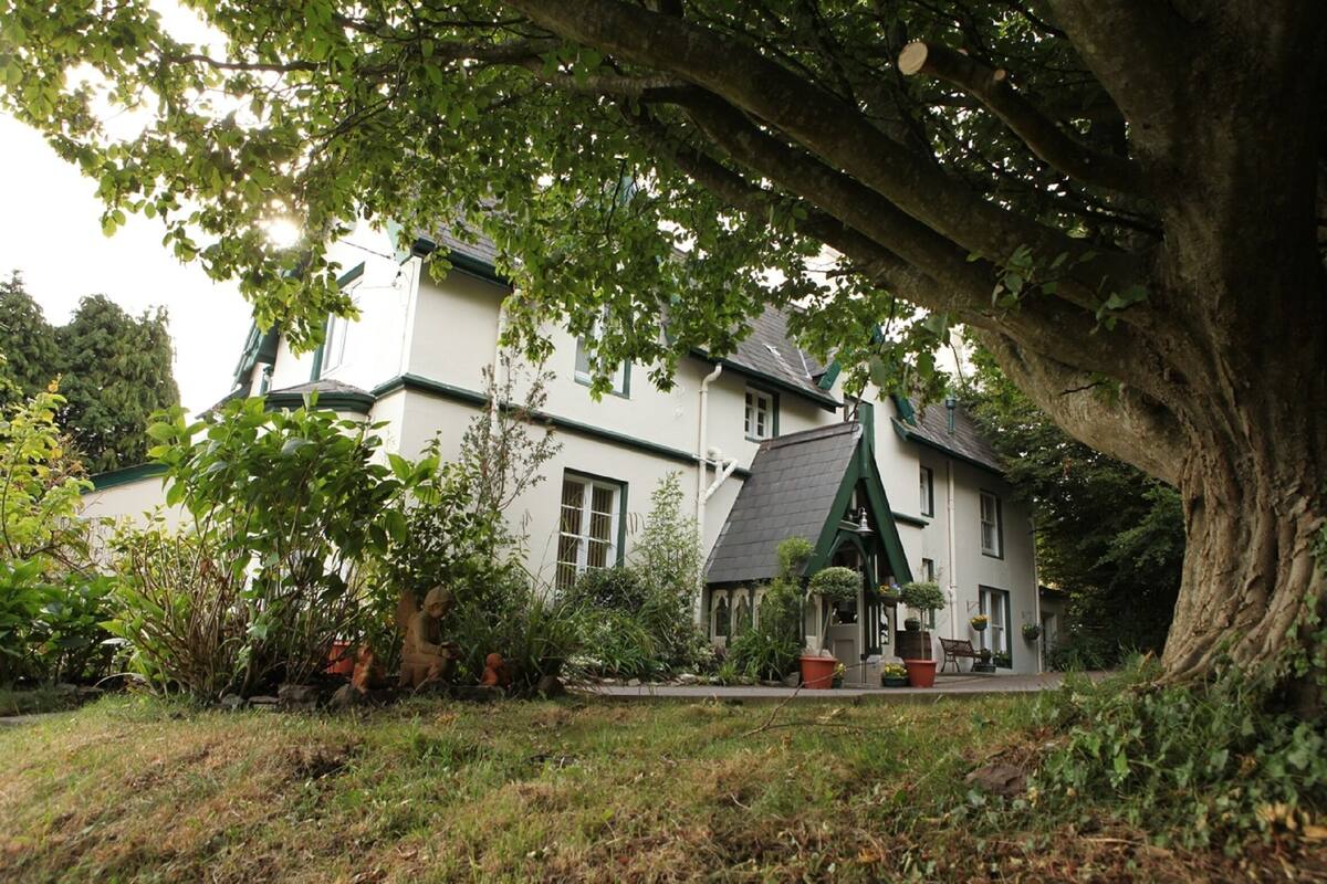 A two-story house with a green roof and white walls, surrounded by lush greenery and trees.