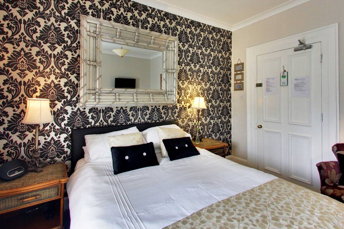 A neatly made bed with white linens and black pillows, a large mirror above it, and patterned wallpaper.
