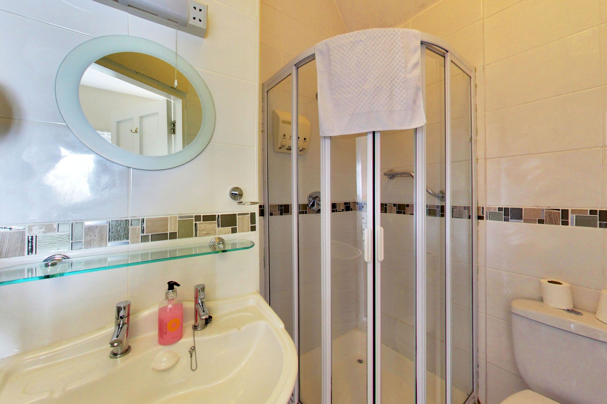 A bathroom with a glass shower enclosure, a glass shelf above the sink, and a toilet with a roll of toilet paper on the wall.