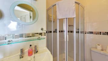 A bathroom with a glass shower enclosure, a glass shelf above the sink, and a toilet with a roll of toilet paper on the wall.