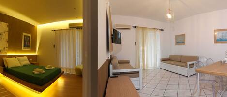 Habitación de hotel con ducha semicircular, cama con ropa de cama verde, sofá, mesa de comedor y zona de cocina.