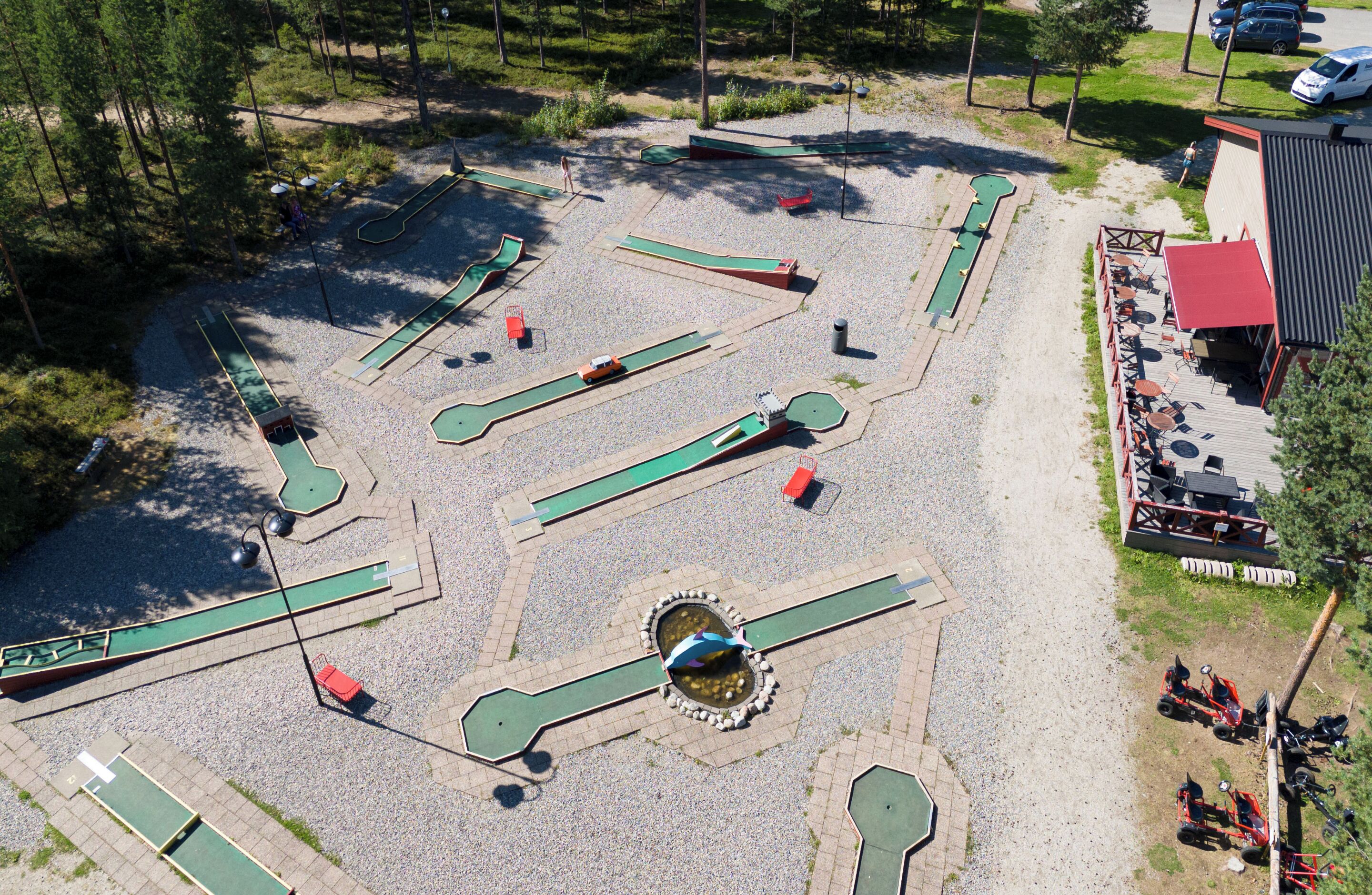 An aerial view of a golf driving range with multiple lanes, a central water hazard, and a building with outdoor seating.