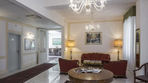 A spacious lobby with a chandelier, red sofas, a round coffee table, and a dining area in the background. - Hotel Villa delle Fonti (Riolo Terme)